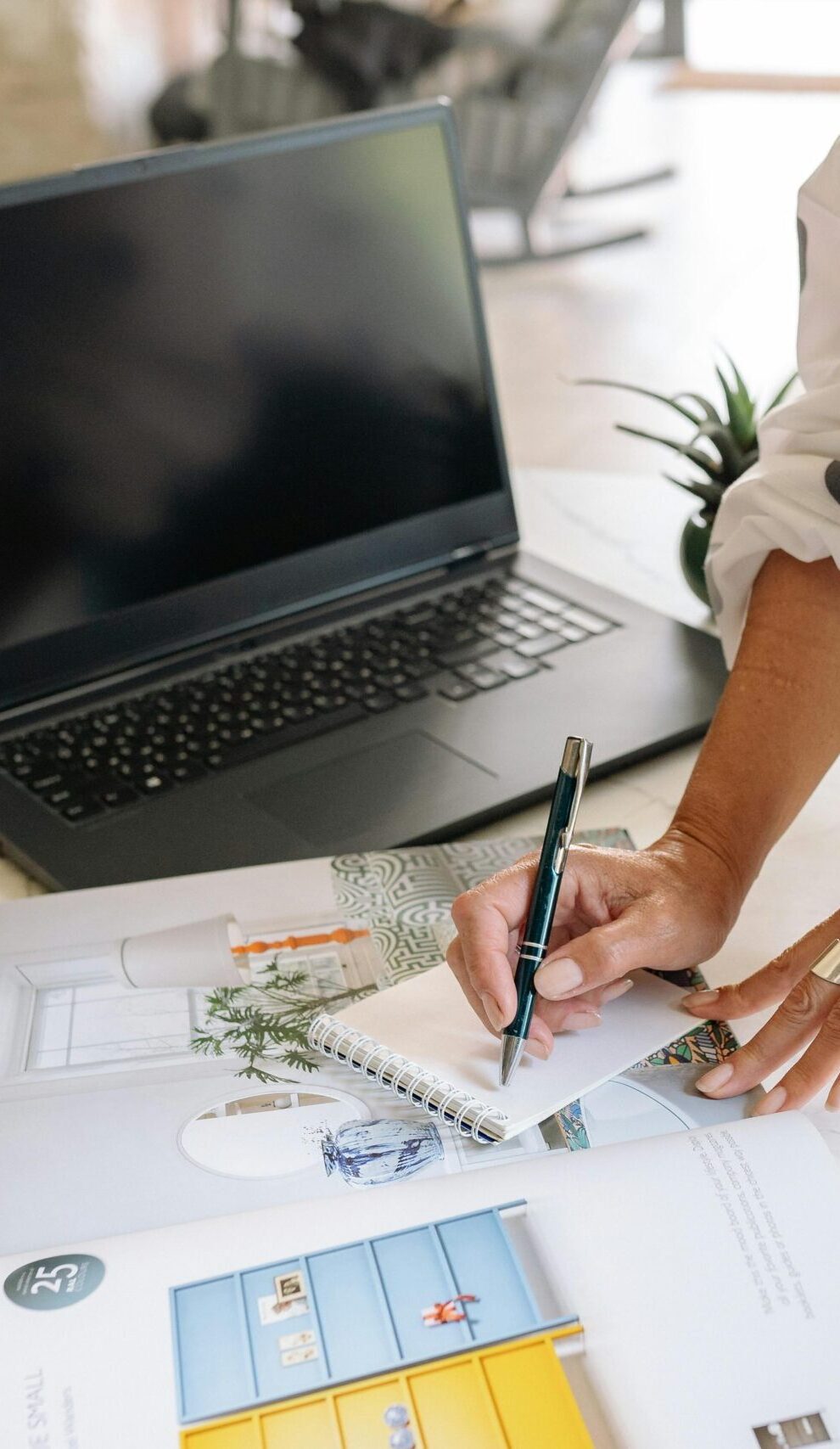 A person writes notes in an office setting with a laptop, magazine, and notepad.